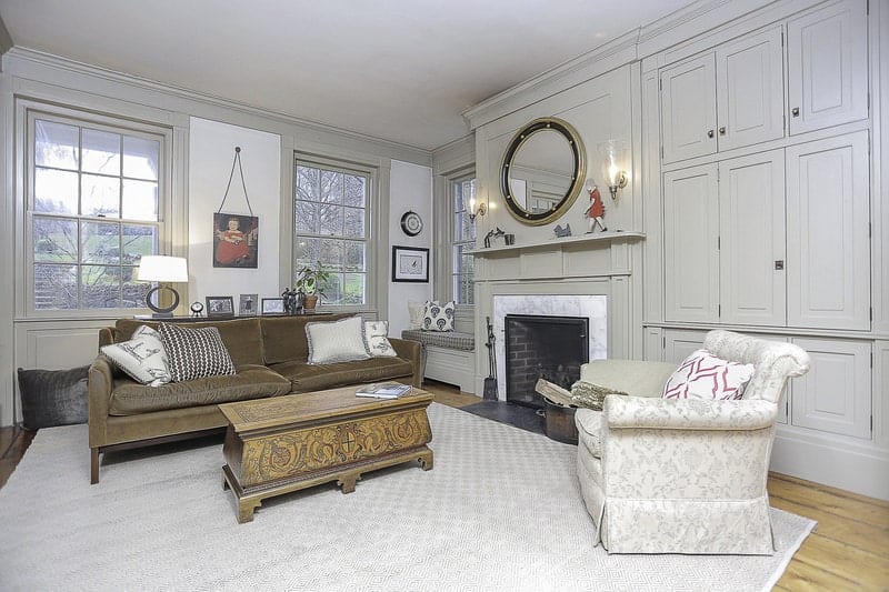 The light gray ceiling and light gray patterned area rug matches with the light gray wooden finish of the walls that has built in cabinets on one side, a fireplace in the middle with a wall-mounted mirror on top and a charming reading nook at the corner by the windows.