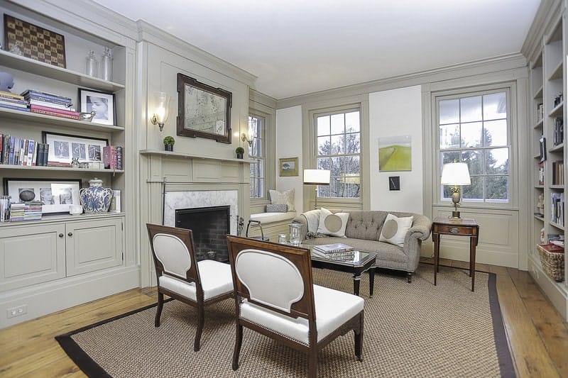 This is a charming Farmhouse-style living room with walls dominated by bookshelves and a fireplace that brings warmth to the light gray tufted sofa and the two wooden chairs with contrasting white cushions facing a glass-top coffee table.