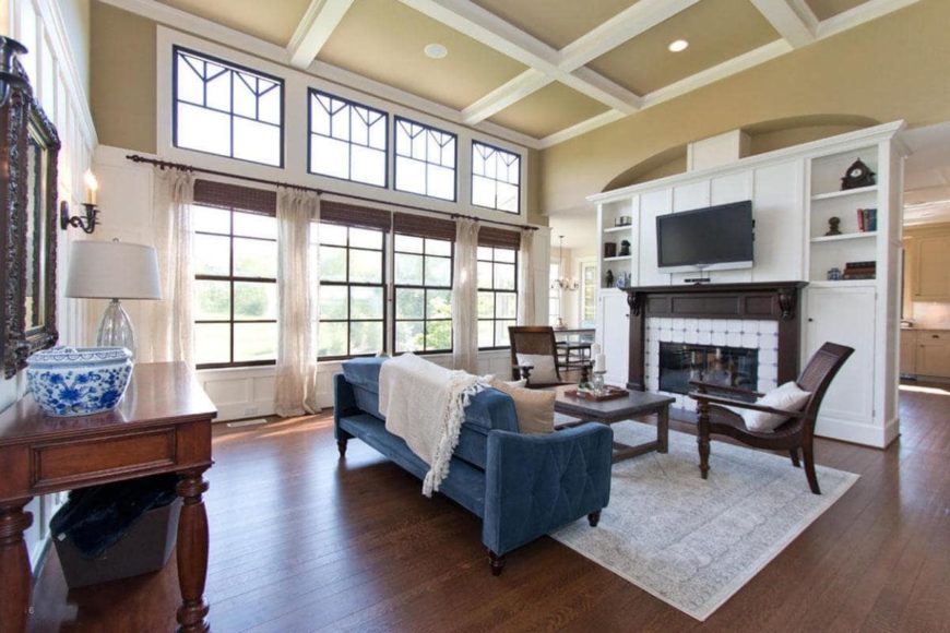 The dark hardwood flooring of this living room is lightened by the tall glass windows that extend all the way up to the beige coffered ceiling that matches the walls contrasted by the dark wooden mantle of the fireplace and the two wooden armchairs.