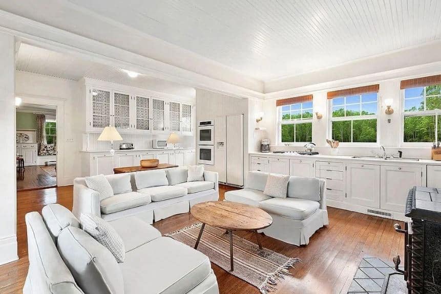 This is a cozy living room that is apparently inside the kitchen. It sits in the middle of the hardwood flooring that is surrounded by kitchen peninsulas with white shaker cabinets and drawers. This matches with the light gray cushioned sofa set surrounding a small elliptical wooden coffee table.
