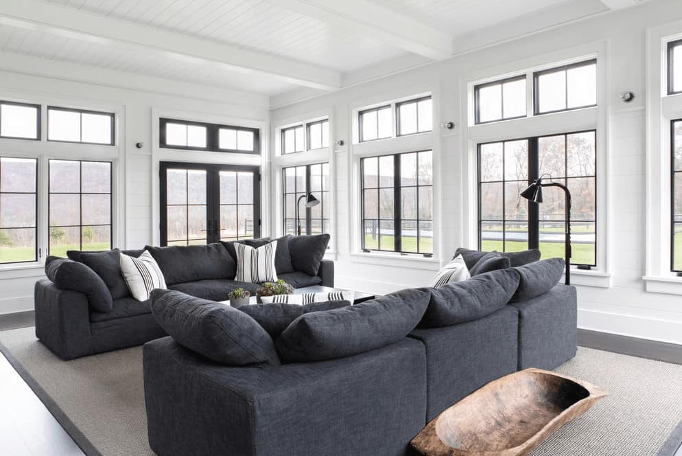 This is a bright Farmhouse-style living room with a white wooden ceiling that has white exposed wooden beams matching with the white walls that bring in an abundance of natural lighting through the surrounding tall windows that lighten up the gray comfortable sofas.