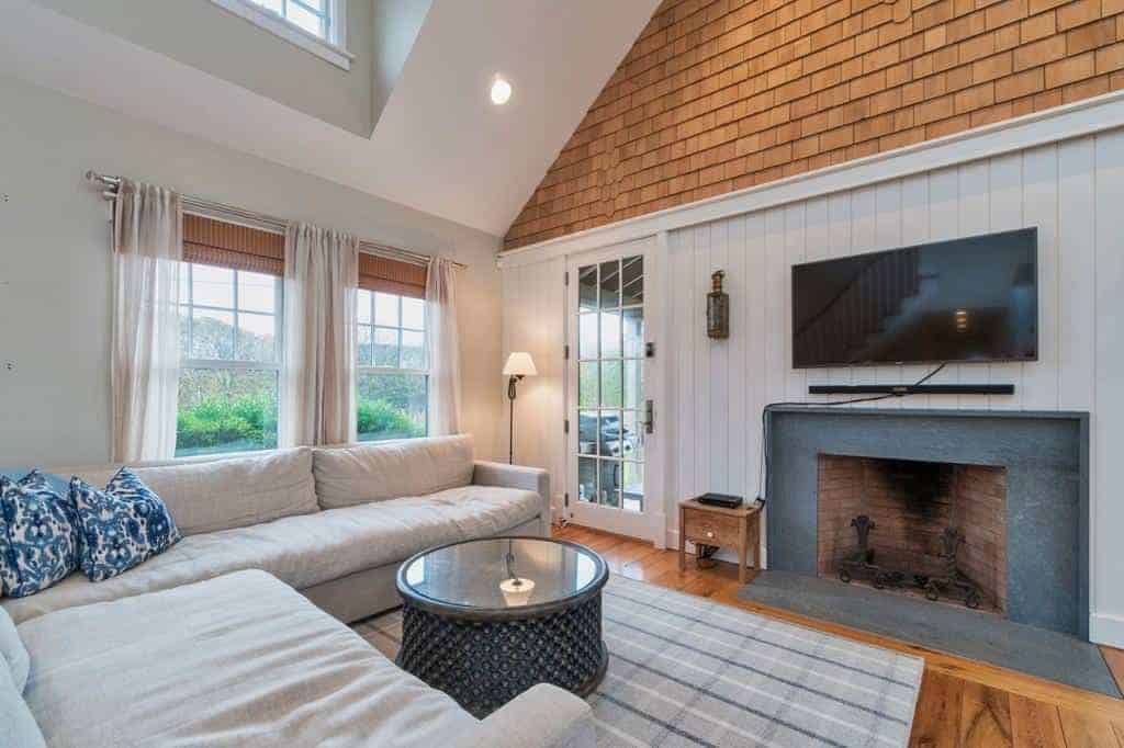 This small and cozy Farmhouse-style living room is brightened by the natural lights coming from the windows and glass door that feature a green landscape outside. This lightens up the gray sectional sofa and the fireplace inlaid with dark gray marble.