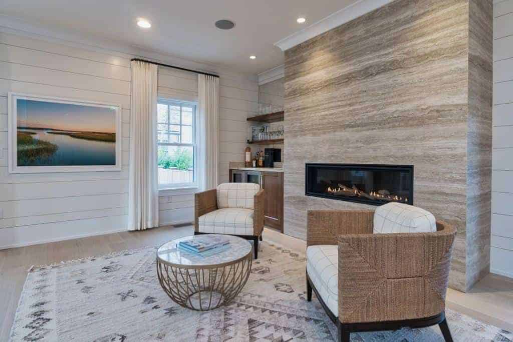 This simple living room has a couple of rustic woven wicker armchairs with white patterned cushions that match the distressed patterned area rug facing a modern glass enclosed fireplace inlaid in a large light gray column in the middle of the armchairs.