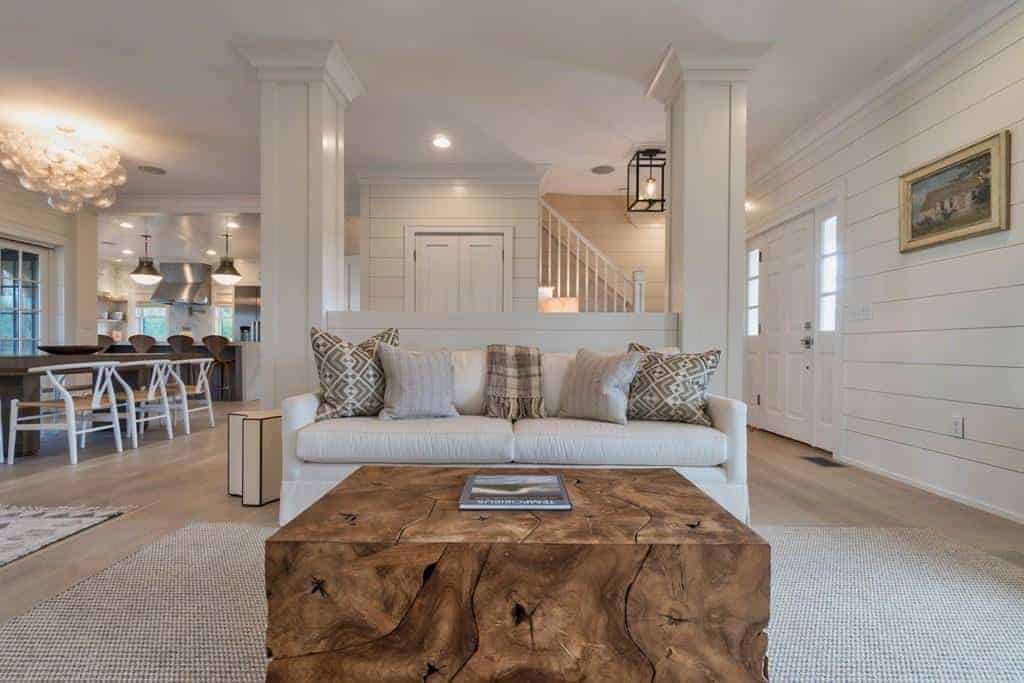 The highlight of this Farmhouse-style living room is the charming wooden coffee table that brings a rustic quality to the light gray area rug over the light hardwood flooring, light gray sofa and light gray walls with a shiplap plank finish.