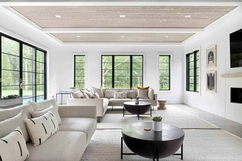 This bright and spacious Farmhouse-style living room has enough space for two light gray sectional sofas that is both paired with decorative round dark coffee tables that stand out against the light gray area rug that is brightened by the surrounding windows.