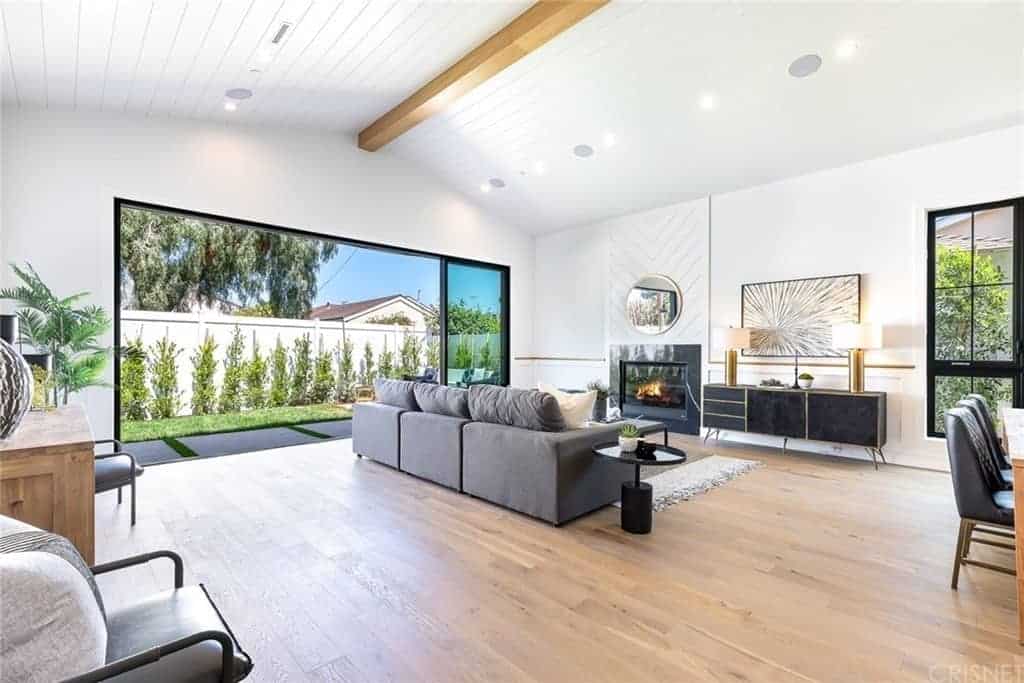 The wide cathedral ceiling with a shiplap plank finish has a single exposed wooden beam running the middle. This is paired with a wide hardwood flooring that is complemented by the gray sectional sofa and the modern fireplace with a dark frame that stands out against the white wall.