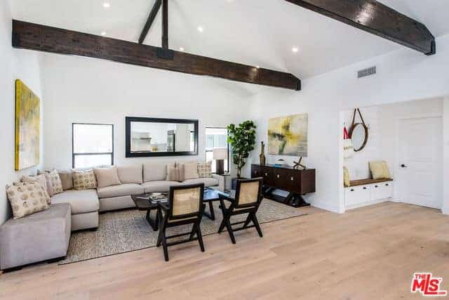 This small living room is placed in an alcove of white walls and two windows with a cathedral ceiling that has exposed wooden beams. The area is dominated by a large L-shaped sectional beige sofa that is contrasted by the dark brown hues of the coffee table, wall-mounted mirror and the wooden armchairs.