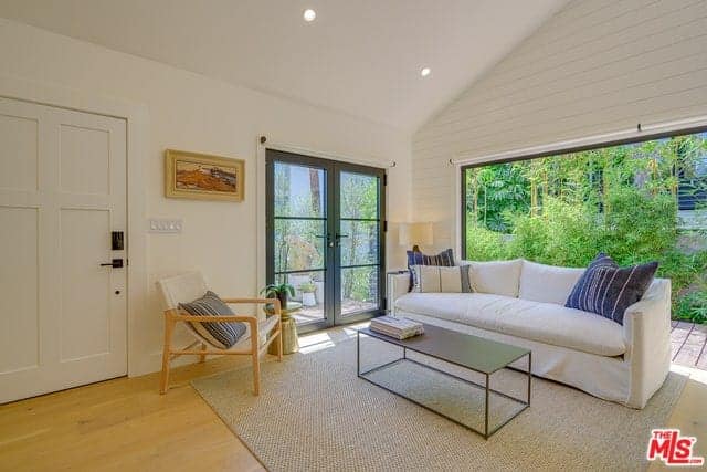 This is a bright and welcoming Farmhouse-style living room that has a white cathedral ceiling paired with white walls that is dominated by a large opening. This showcases the landscaping outside that provides a lush green background for the simple white sofa on a woven area rug.