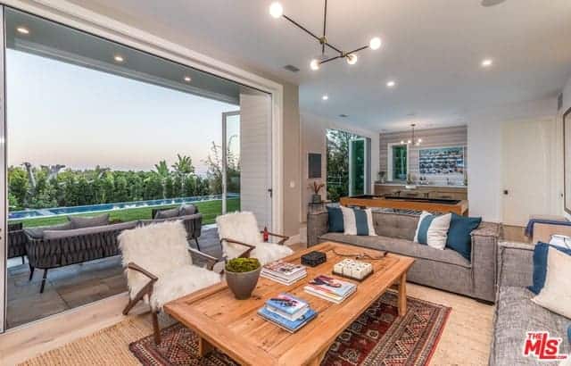 A wonderful view of the backyard pool provides a nice background for the living room that has a light hardwood flooring topped with a colorful patterned area rug under the wooden coffee table that is surrounded by a couple of gray sofas and a couple of wooden armchairs with furry cushions.