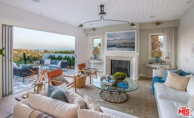 This Farmhouse-style living room is brightened by the large wall that opens up due to the folding glass doors. This brings in an abundance of natural lighting and a wonderful view for those who are lounging on the comfortable white sectional sofa warmed by the white fireplace.