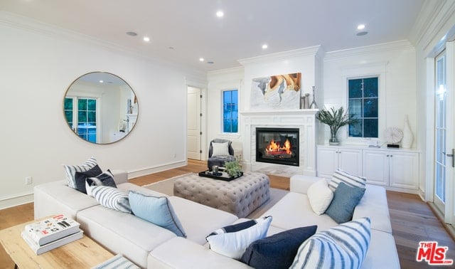 The bright white walls matches with the bright white ceiling that is augmented by recessed lights that shine down on the white mantle of the fireplace that is topped with a colorful painting across from the white L-shaped sectional sofa and its light gray tufted cushioned coffee table.