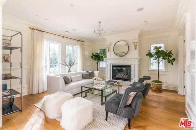 This bright living room has a hardwood flooring that is covered by a light gray patterned area rug. This complements the white cushioned sofa and the pair of cushioned tufted gray chairs across from it beside a couple of white furry ottomans.