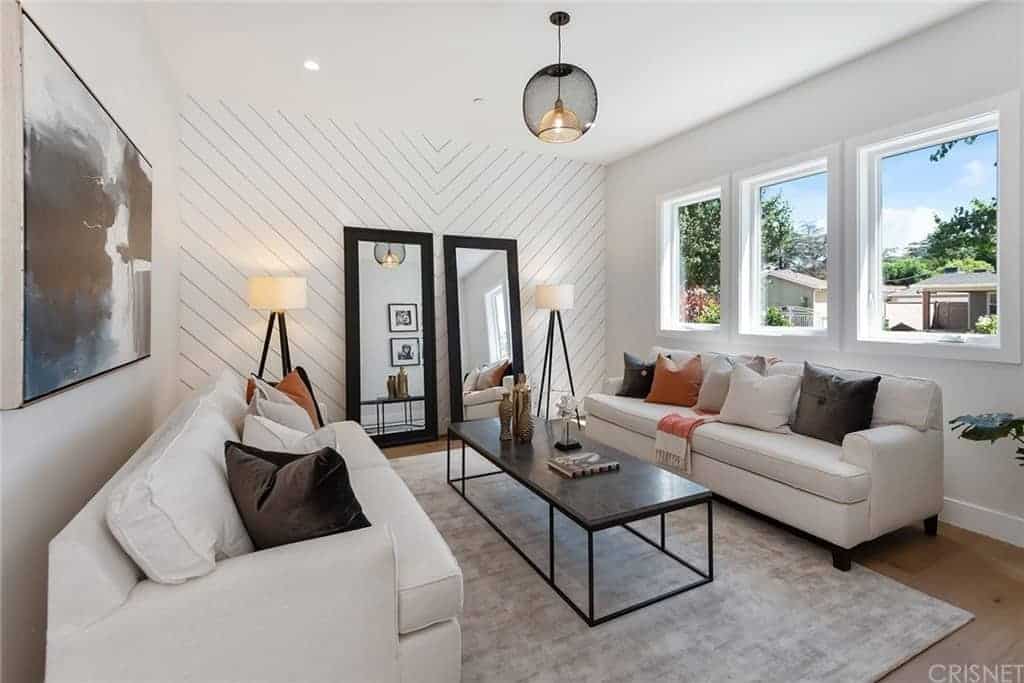 The white wall on the far side has a patterned plank finish and adorned with a couple of tall mirrors flanked by standing lamps. This sets a nice background for the couple of cushioned sofas and the narrow dark coffee table in the middle.