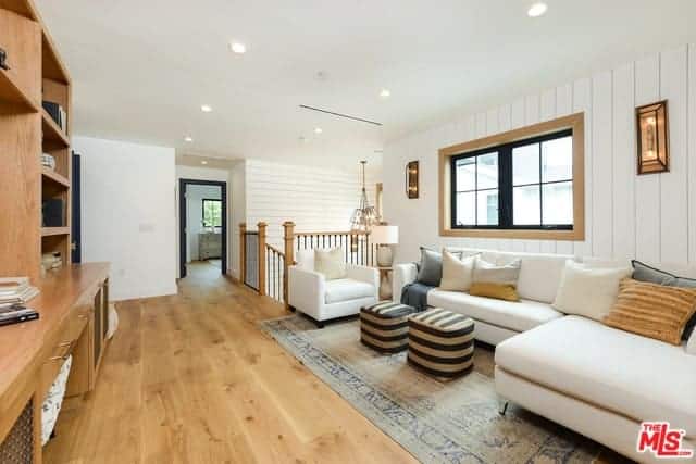 This is a second floor living room right beside the staircase railings. It is against a white wall with a plank finish adorned with a couple of wall lamps flanking the window above the L-shaped sectional sofa facing two striped ottomans.