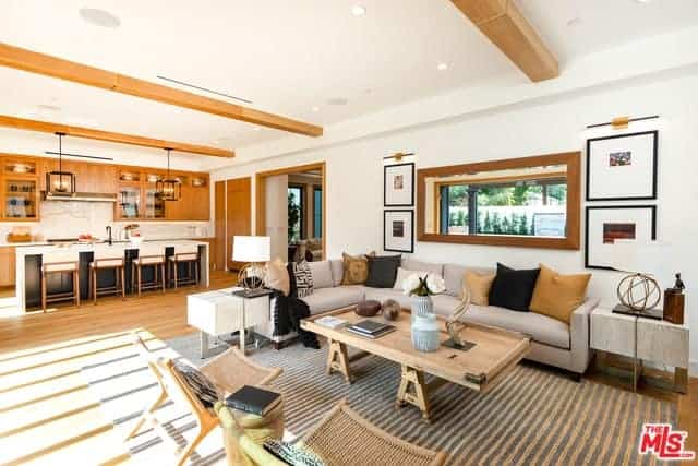 Patches of sunlight are casting down on the light gray patterned area rug over the hardwood flooring. This is matched with the wooden coffee table that is facing the large L-shaped sectional couch with colorful pillows and adorned with a large mirror mounted on the white wall above.