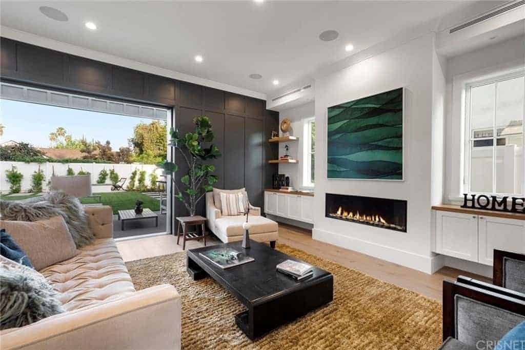 The modern fireplace that is built into the white wall is adorned with a large green painting mounted above it. This looks over a unique black wooden coffee table that complements the light pink cushioned sofa and the brown rustic area rug.