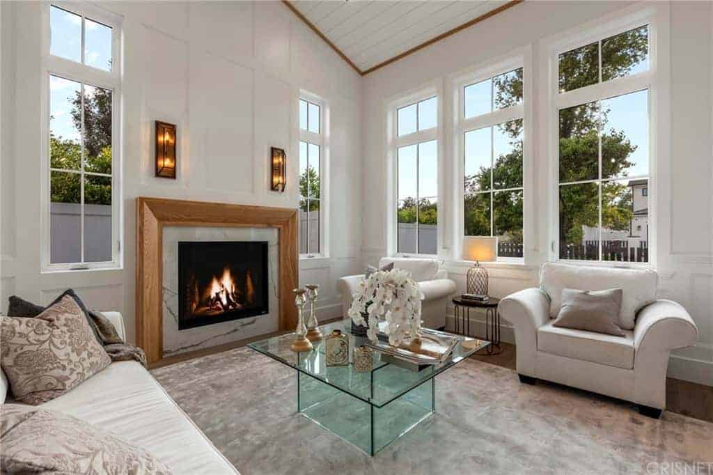 The glass coffee table stands in the middle of the gray area rug flanked by the light gray comfortable sofa and cushioned armchairs brightened by the surrounding tall windows and warmed by the fireplace built into the wall with a wooden mantle.
