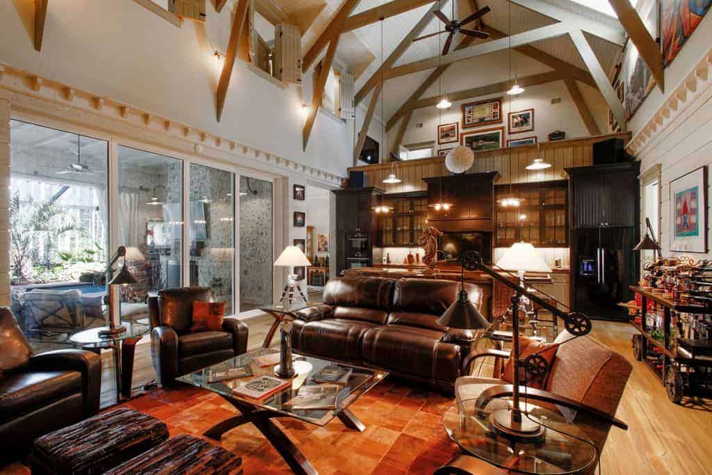 This living room area is part of a large room with a high cathedral ceiling that also houses the kitchen on the far side. The living room has brown leather cushioned sofas and armchairs that complement the rust-colored area rug underneath the glass-top coffee table.