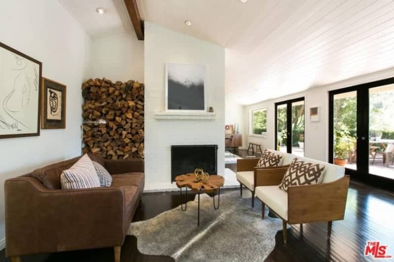 This Farmhouse-style living room has a dark hardwood flooring that is contrasted by the wall of the fireplace that also houses the fire wood on the side. This is paired with a brown leather sofa and couple of wooden armchairs with white cushions.