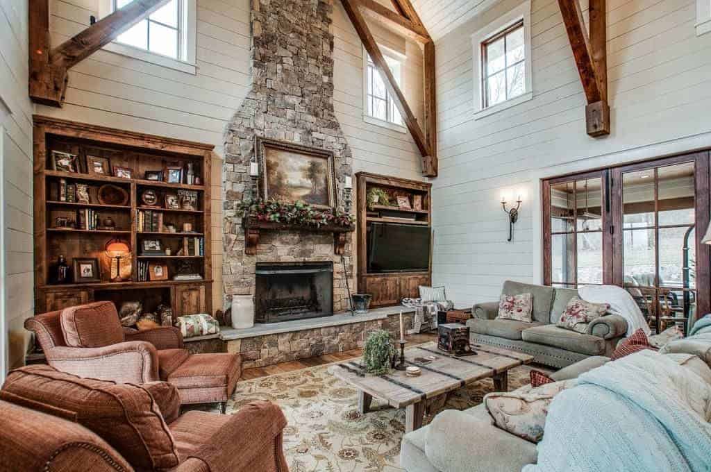 The brown, gray and light gray cushioned sofas and armchairs are all facing a rustic wooden coffee table across from a large wall that is dominated by the large stone column housing the fireplace that is flanked on both side by wooden shelves.