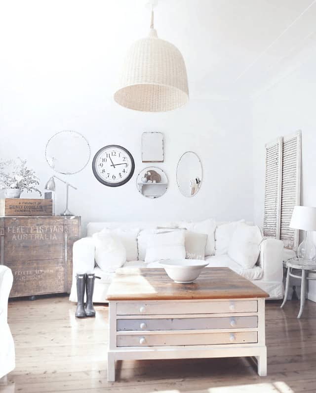 The bright living room features a wooden coffee table with storage lighted by a wicker dome pendant. It is decorated with a wall clock and various shaped mirrors mounted above the white sofa.