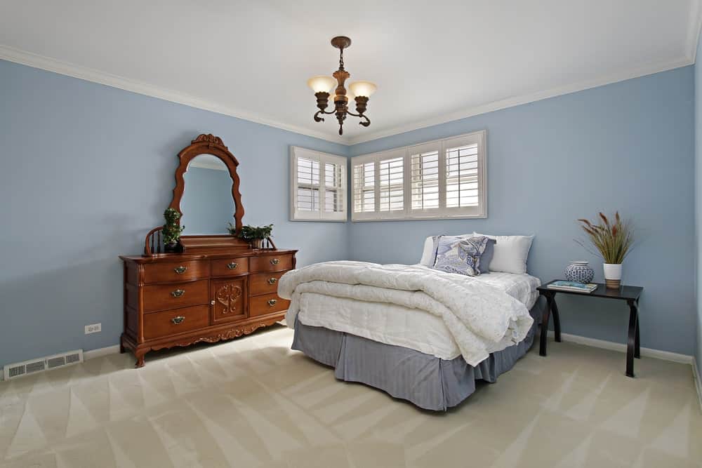 A vintage chandelier hangs over the skirted bed that's accompanied by a black nightstand and wooden dresser placed against the muted blue walls.