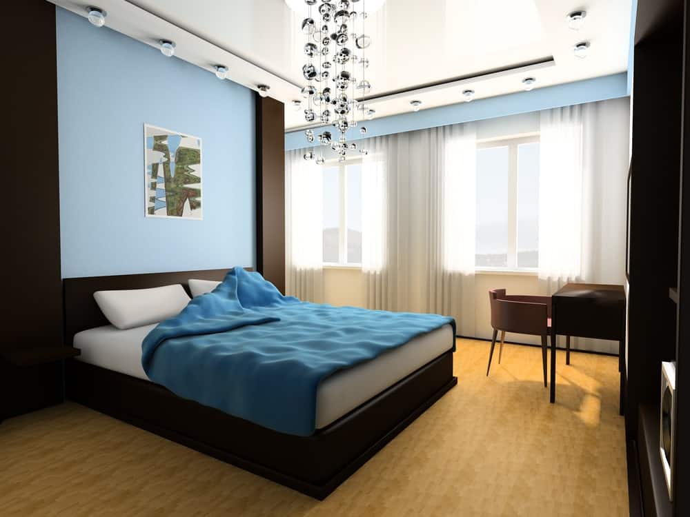 Modern primary bedroom illuminated by a glass bubble chandelier and globe flush lights fitted on the tray ceiling. It has a sleek black bed and wooden desk paired with a round back chair.