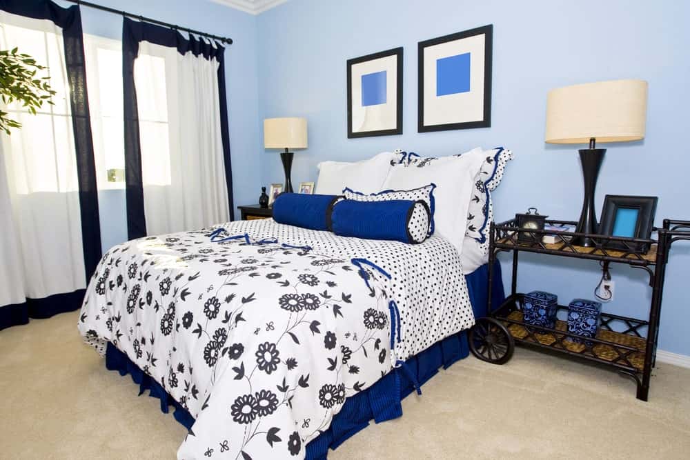 Sleek drum table lamps sit on black nightstands flanking a blue skirted bed dressed in dotted and floral bedding. It is accented with framed wall arts mounted on the light blue wall.