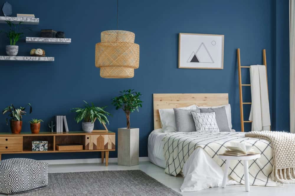 Fresh bedroom offers a wicker chandelier and light wood bed with a ladder on its side that serves as a rack. It includes marble floating shelves and a patterned ottoman that sits on the gray area rug.