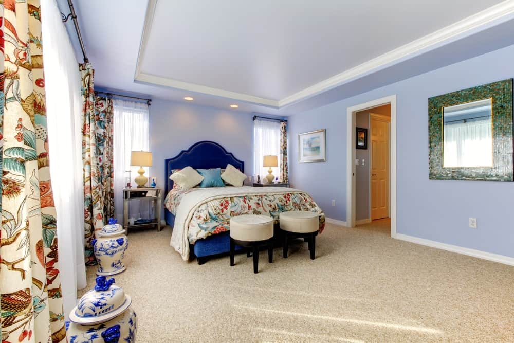 Printed draperies complement the bedding in this primary bedroom showcasing ceramic vases and a green mosaic mirror mounted on the soft blue wall.