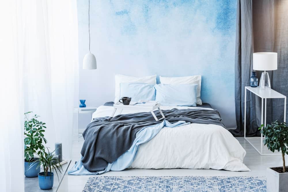 Green potted plants create a refreshing ambiance in this airy primary bedroom with sleek white tables and comfy bed illuminated by a dome pendant light and glass table lamp.