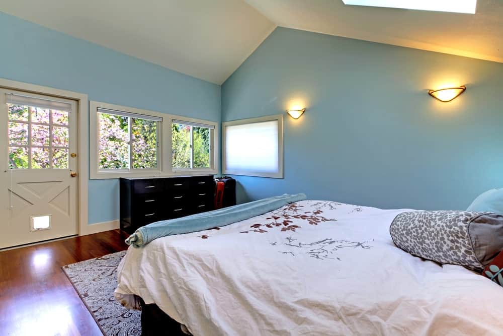 This bedroom boasts dark wood dresser and a comfy bed lighted by glass sconces that are mounted on the sky blue wall. It has a vaulted ceiling and rich hardwood flooring topped by a patterned area rug.