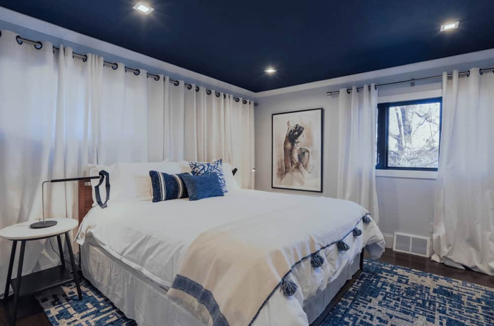 A blue printed rug lays on the dark hardwood flooring in this primary bedroom featuring a gorgeous painting and comfy bed illuminated by recessed ceiling lights.