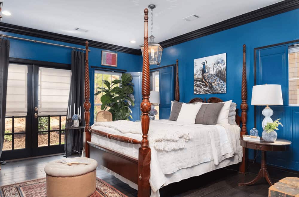 Classic blue bedroom decorated with a lovely peacock artwork and lantern pendant light that hung over the four poster bed accompanied by white table lamps and round ottoman.
