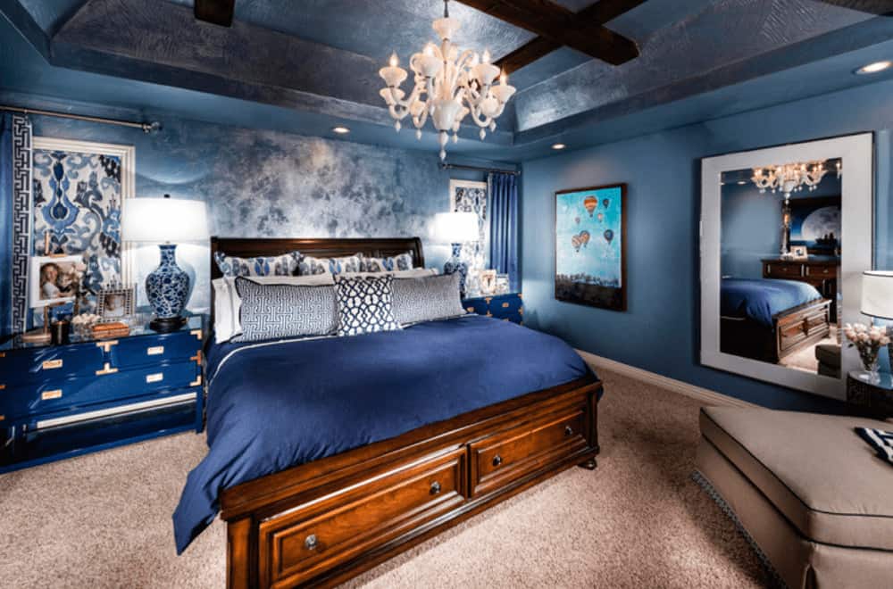 A gorgeous white chandelier illuminates this primary bedroom boasting a wooden bed and ceramic table lamps that sit on the blue nightstands.