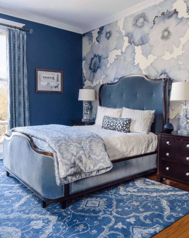 Clad in floral wallpaper, this blue bedroom offers a tufted wingback chair that sits on a patterned area rug. It is accompanied by dark wood nightstands and glass table lamps.