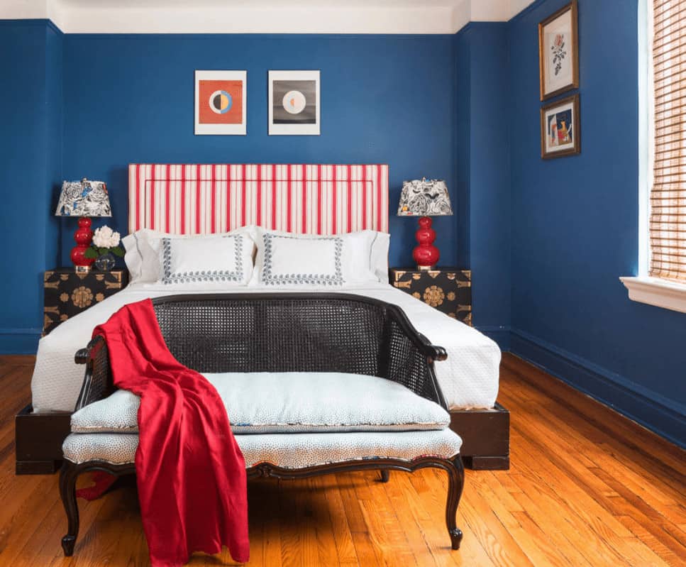 A wicker cushioned bench sits in front of the wooden bed with red striped headboard flanked by charming table lamps and floral nightstands.