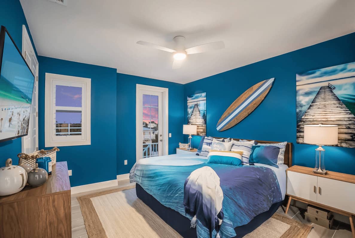 Beach style bedroom decorated with gorgeous artworks and a decorative surfboard mounted above the comfy bed. It has a jute area rug and wooden nightstands topped with glass table lamps.
