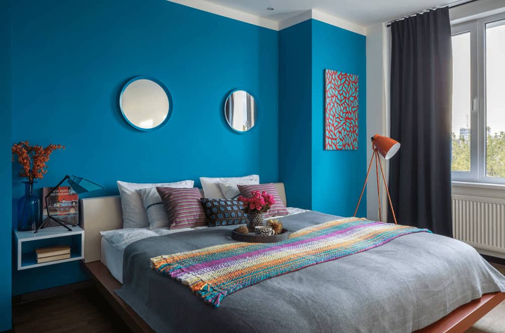Blue bedroom showcases a lovely artwork and multi-color knitted throw that lays on the cozy bed. It includes round mirrors and floating shelf topped with stylish lamp and translucent vase.