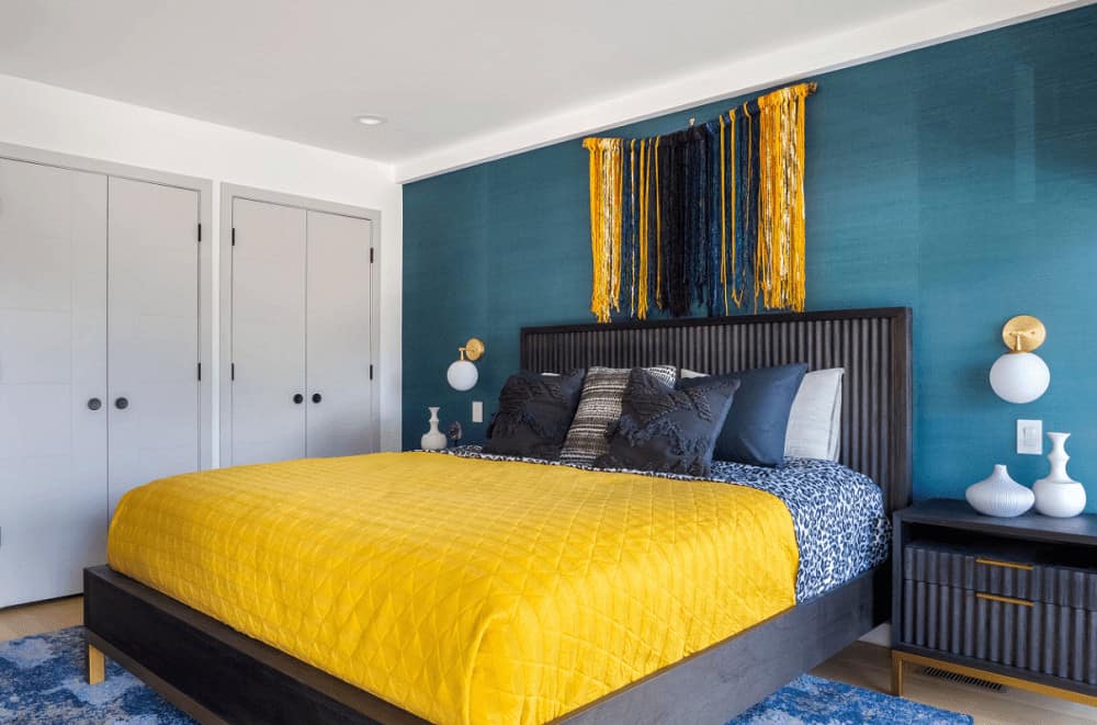Contemporary bedroom decorated with a gorgeous yarn tapestry that hung above the wooden bed dressed in yellow bedding. It has globe sconces and white vases that sit on dark wood nightstands.