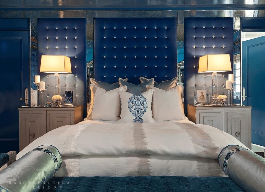 Luxury primary bedroom with a classy bed and wooden nightstands placed against the mirrored wall that's fitted with blue tufted panels.