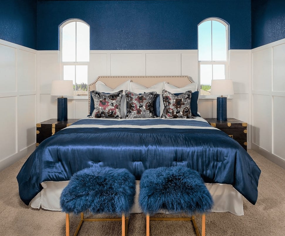 Transitional bedroom offers a gorgeous bed surrounded by wainscoted walls and arched windows. It includes faux fur stools and blue table lamps that sit on dark wood nightstands.