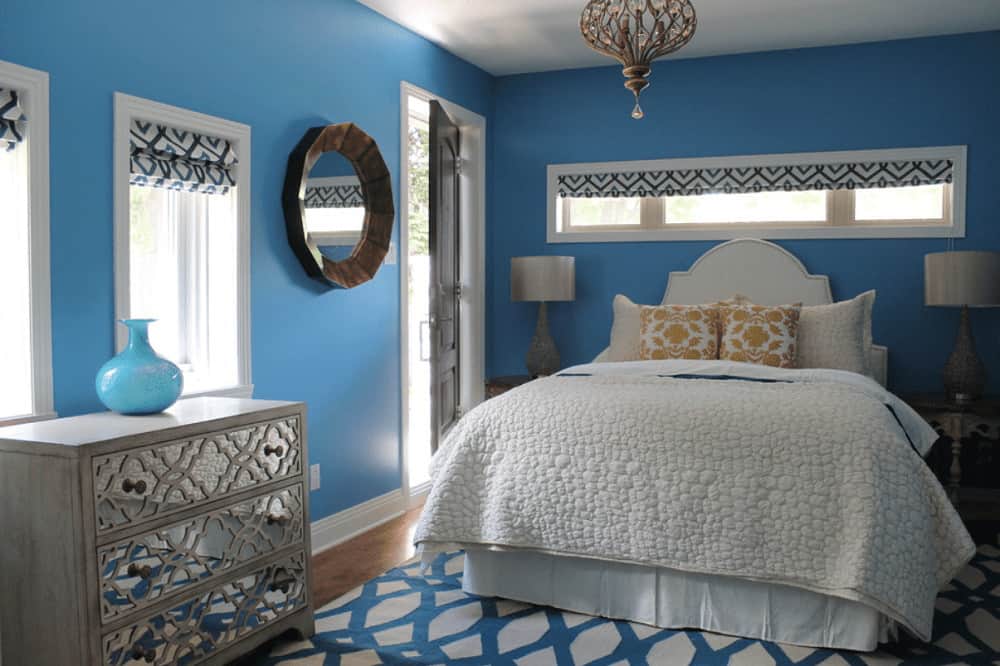 Fabulous bedroom with a charming chandelier and round mirror mounted on the blue wall. It includes a skirted bed and mirrored drawer chest topped with a blue vase.