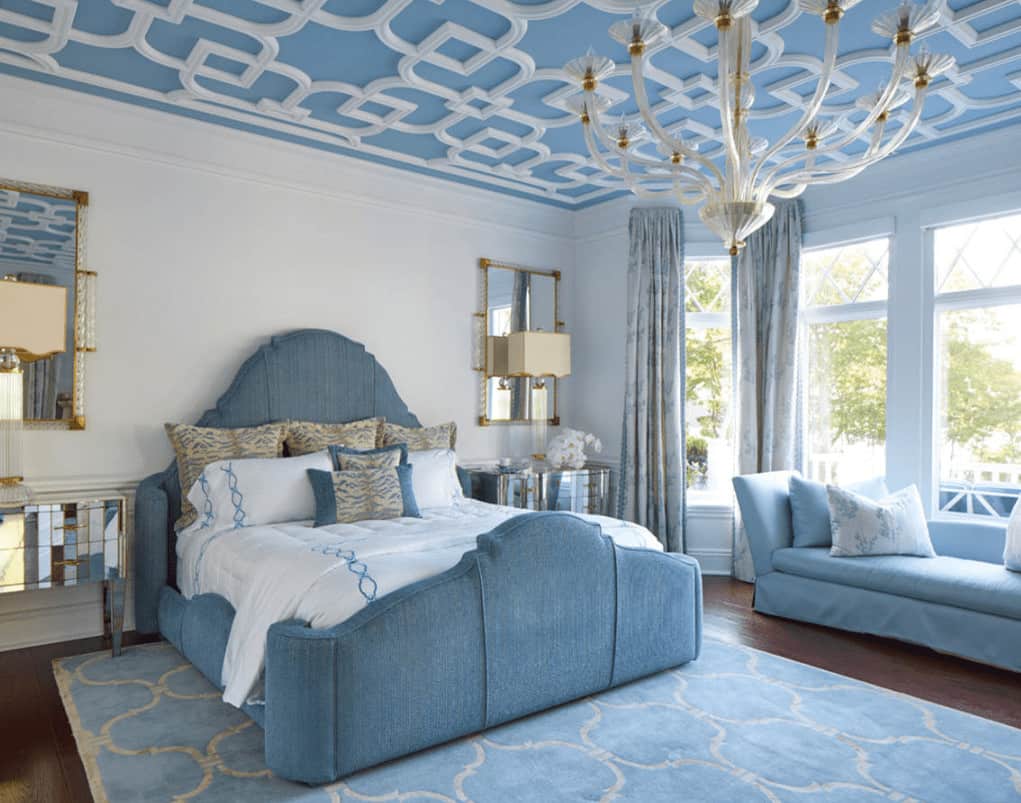 Stylish bedroom with mirrored nightstands and a blue upholstered bed accompanied by a skirted seat. It is illuminated by an oversized white chandelier that hung from the ornate ceiling.