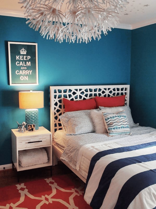 A blue table lamp sits on the light wood nightstand in this bedroom with a framed artwork and white ornate bed over a red patterned rug.