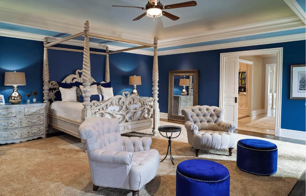 Beige tufted armchairs paired with blue velvet ottomans sit in front of the four poster bed complementing the distressed nightstands topped with chrome table lamps.