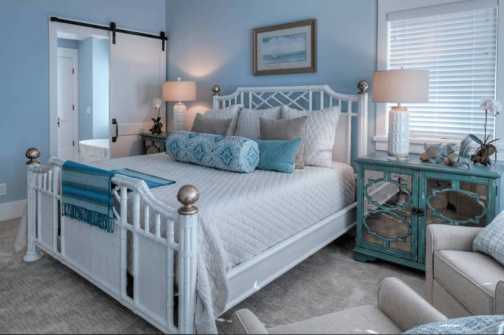 Beach style bedroom with white wicker bed and mirrored nightstands in distressed blue. It includes drum table lamps and gray armchairs that blend in with the carpet flooring.