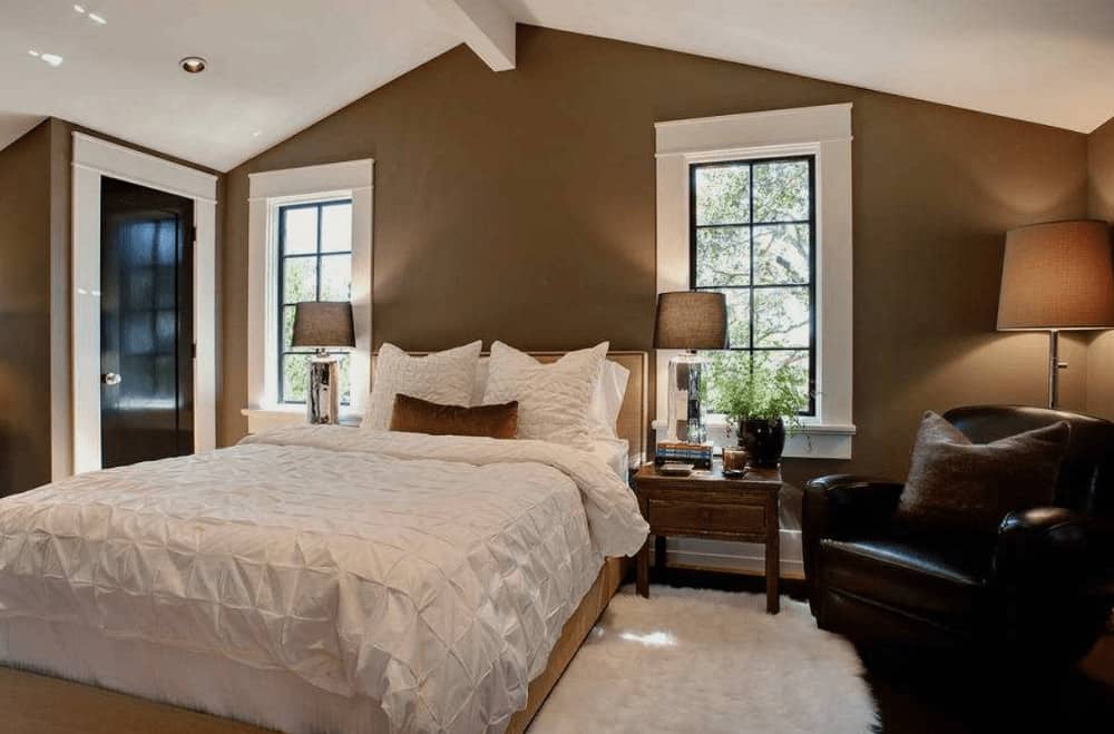 Natural wood nightstands and mirrored table lamps flank a cozy bed in this brown primary bedroom boasting a white shaggy rug and black leather armchair lighted by a traditional floor lamp.