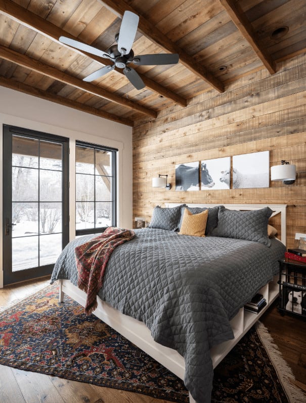 Cozy primary bedroom with multi-panel wall art and white bed on a tasseled rug wrapped in a gray quilt. It has black framed windows and wood plank ceiling mounted with a fan.