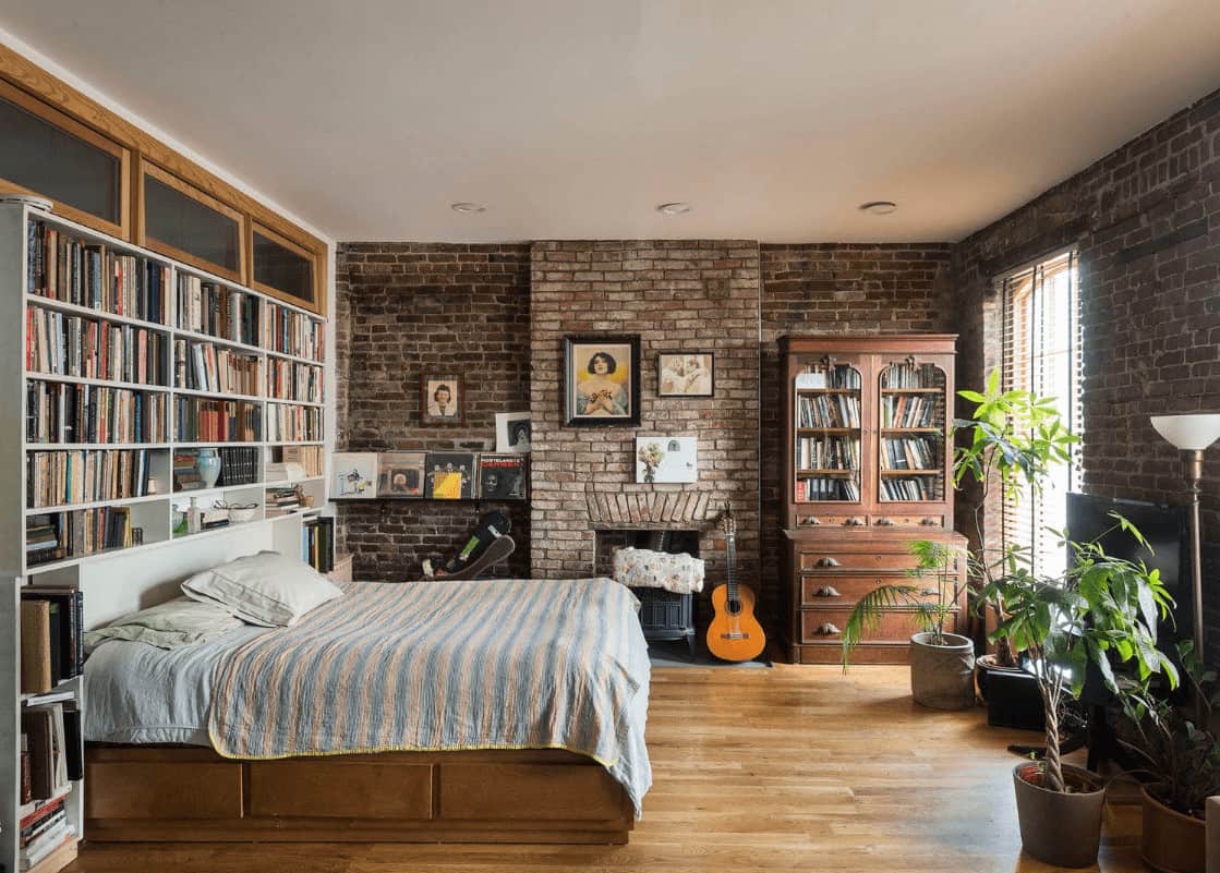 Green potted plants create a refreshing ambiance in this primary bedroom showcasing built-in bookshelves and a fireplace concealed in the brick walls.